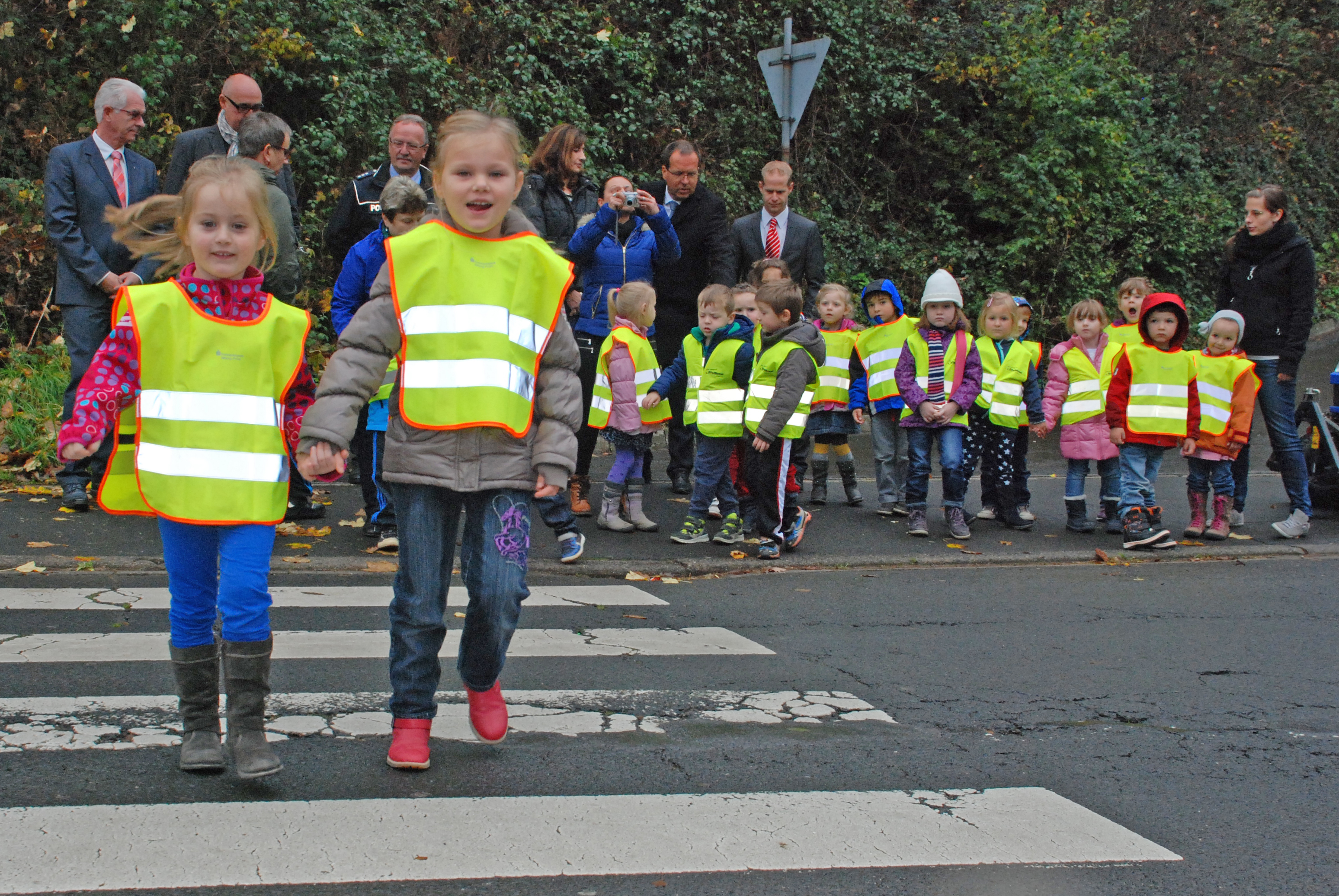 Zwei Kinder mit gelben Warnwesten gehen über einen Zebrastreifen. Weitere Kinder warten auf dem Bürgersteig.